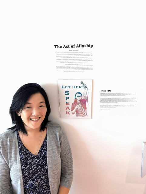 Joy standing in front of her portrait of Zoey Zephyr and the description of the exhibition, The Act of Allyship.