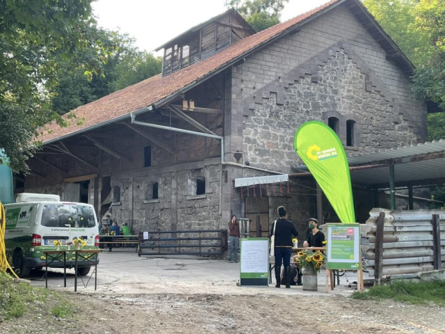 Ein großer, rustikaler Scheunenbau aus Naturstein und Holz steht am Rand eines bewaldeten Geländes. Vor dem Gebäude befinden sich mehrere Menschen an Tischen und Infoständen. Auffällig ist eine grüne Fahne mit dem Logo von Bündnis 90/Die Grünen. Links steht ein Lieferwagen, daneben ein kleiner Tisch mit Sonnenblumen. Die Atmosphäre wirkt wie ein sommerliches Treffen oder eine Veranstaltung auf einem Bauernhof oder Kulturgelände.