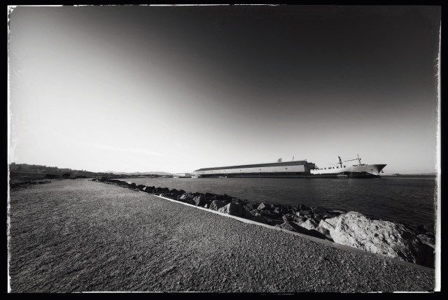 An ultra wide photo of a packed dirt path on the edge of an inlet on San Francisco's eastern shoreline. 

Across the inlet: a long, low industrial shed (a giant recycling facility and popular perch for seagulls). On the right: a ship pointing out into the San Francisco Bay. On the left: glare in the sky from the late afternoon sun.

Harsh. Stark. Desolate. Lacking people. Lacking trees. Also monochrome.