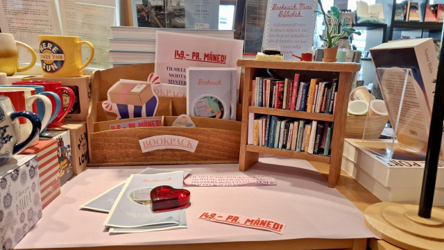 Short version: book display with tiny books. Fuller description: Close-up of a hand-made display at family-run independent bookshop Kristian F Møller in Aarhus, Denmark. It advertises the Bookpack monthly subscription at 149 kroner per month – great value in a country where one book usually costs about 250 kr or 300 kr (€35 or €40). Surrounded by colourful coffee mugs and books and gift cards,there is a tiny wooden bookshelf about the size of an average paperback. On each of its two shelves, about twenty teeny colourful matchbox-sized ‘books’, each with the author name clear on the spine. The covers are faithful to the real book covers. Author names include Helle Helle. RE Kuang. Patti Smith. Bonnie Garmus. Murakami. Katrine Marie Guldager. Bernadine Evaristo. Douglas Stewart. Thomas Mann. Ann Patchett. Chimamanda Ngozi Adichie. 