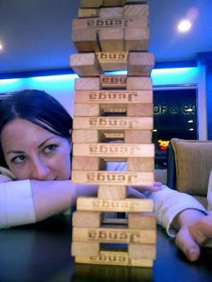 A cute, smiling, dark-haired person, with their head at table-height, looking up at a tall Jenga tower.