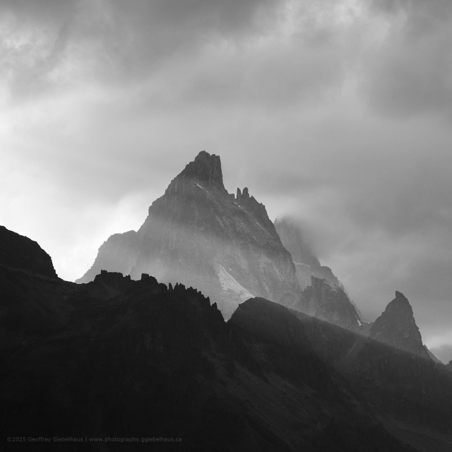 A dramatic jagged mountain peak emerges through swirling mist and brooding storm clouds in this moody black and white Canadian Rockies landscape photograph, its dark rocky summit rising sharply into a luminous break in the grey sky with two distinctive rock spires at its crown that bear a striking resemblance to a hand making a peace sign. The monochrome treatment deepens the contrast between the inky silhouetted foreground ridgelines and the bright, cloud-diffused light illuminating the central peak, amplifying the raw power and theatrical atmosphere of the scene. This striking black and white mountain nature photograph proves that the wilderness has a sense of humour, hiding playful moments of joy within its most dramatic and imposing landscapes.