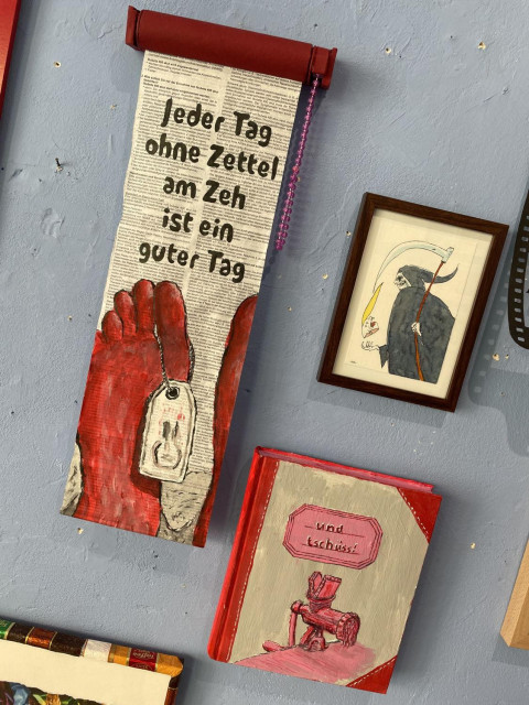 an einer Grauen Wand hängen drei Kunstwerke. Eines zeigt einen auf Zeitung gemalten Fuß von unten an dem ein Zetetl hängt. Darüber steht geschrieben 'jeder Tag ohne Zettel am Fuß ist ein guter Tag'