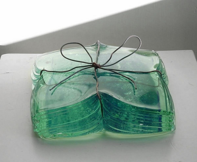A stack of square, translucent green glass plates or coasters resting on a white surface. The stack is tied together with a thin dark wire that forms a simple bow on top. Soft light hits the glass from the left, creating subtle shadows and highlights.