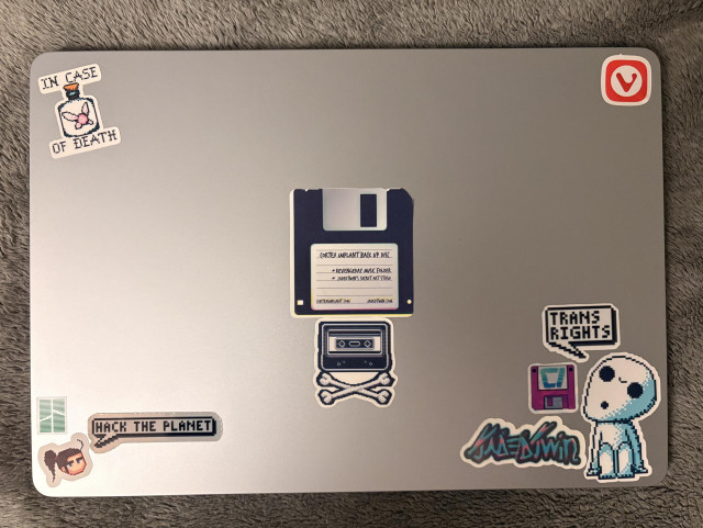 A MacBook Air, with a number of stickers from jadedtwin and Cortex Implant, including quote bubbles for “Hack the Planet!” and “Trans Rights”. A Vivaldi sticker is alao included. 

The beginning of the stickering has begun.