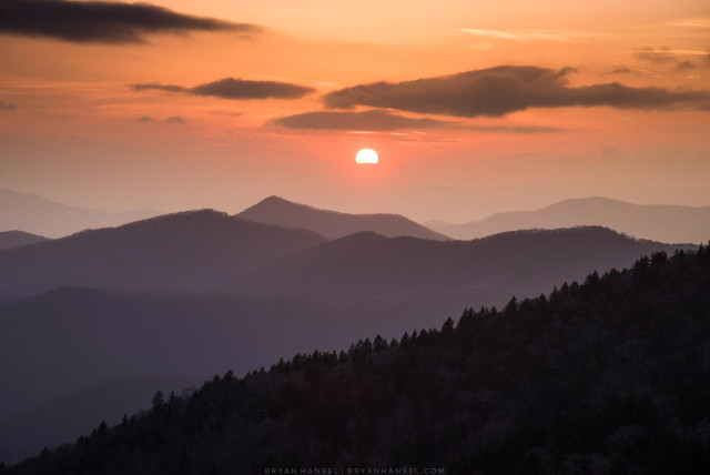 Under an orange sky the sun disk disappears behind distant clouds covering distant mountain ridges and the mountain ridges come forward in the shot -- layer after layer. 