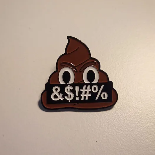 Photo of an enamel pin from the EFF of the enshittification artwork (the poop emoji with its mouth covered by grawlix).