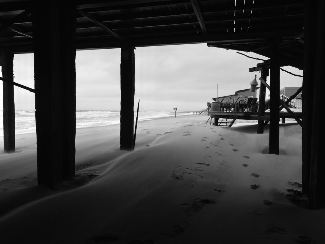 ein schwarz-weiß-foto zeigt den blick von unterhalb einer hölzernen terrasse oder eines strandcafés auf einen strand. im vordergrund sind dunkle, schattige stützpfeiler zu sehen, während sich auf dem sandboden sanfte verwehungen und eine spur von fußabdrücken abzeichnen. im hintergrund erstreckt sich die küstenlinie mit einer brandung unter einem bewölkten himmel. rechts im bild ist ein anderes strandhaus erkennbar. es wirkt kalt, windig und ungemütlich.