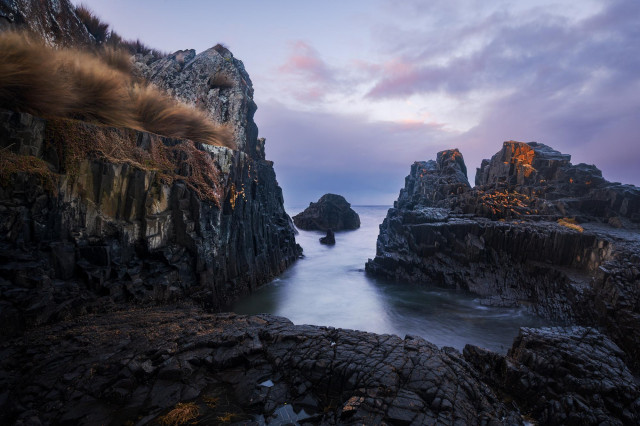 sunset seascape with volcanic rock cliff on the left and a rock platform on the right. The rocks are covered with orange lichen and yellow-orange patches of grass. The composition forms a corridor between to sections leading to another submerged rock in the middle. The sky is pale lavender and blue.