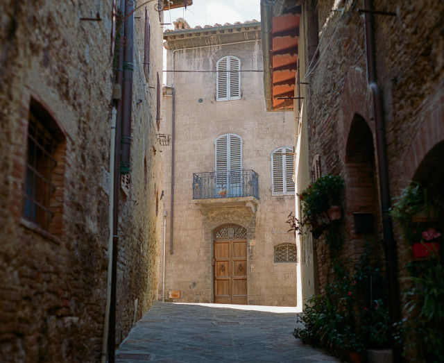 A dark medieval street with a visible incline, green potted plants on one side, a building illuminated by light at the end of the street with a wooden double door and a balcony above it. All the windows are covered in light blue shutters.