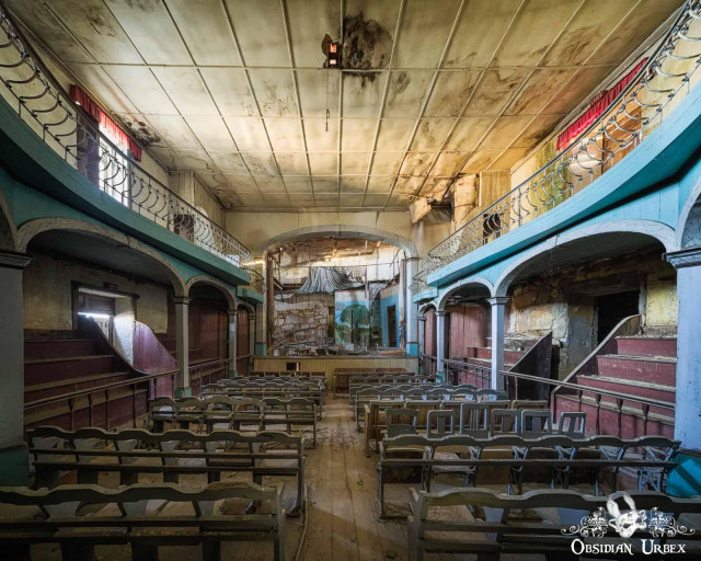 An abandoned theater auditorium features rows of empty wooden chairs facing a decaying stage, with balconies on either side.