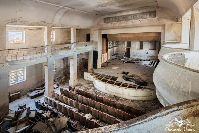 A high-angle view of a crumbling, abandoned theater with rows of broken seats and a decayed stage.