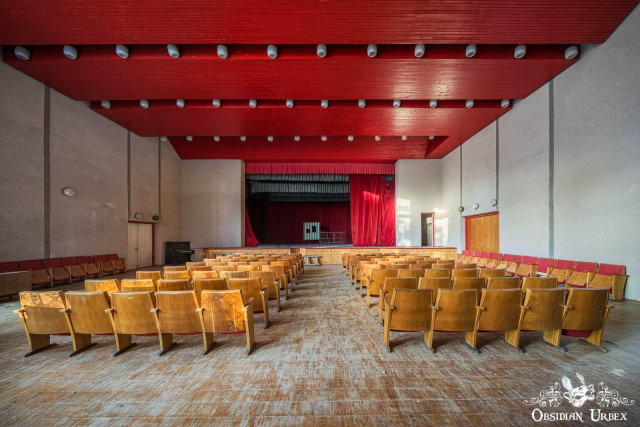 Rows of empty wooden chairs face a dusty stage with red curtains under a vibrant red ceiling in an abandoned theater.