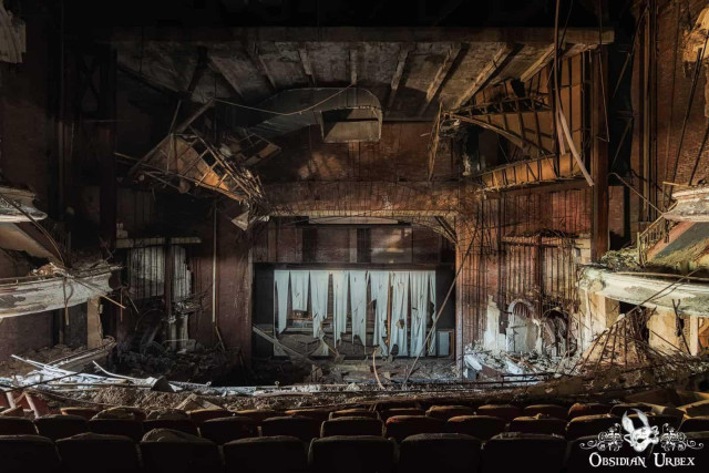 A wide shot of a decaying abandoned theatre with collapsing ceilings, torn stage curtains, and rows of empty seats.