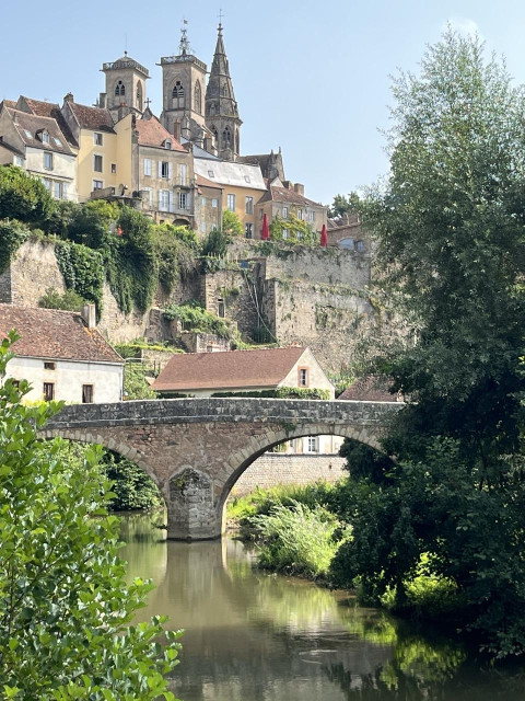 Un vieux petit pont sur un petit cours d'eau en bas de Semur-en-Auxois. La cathédrale en hauteur, des arbres et un ciel dégagé. 