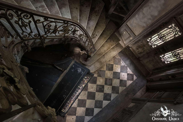 View from above of a grand, dusty staircase curving around a black piano on a checkered floor in an abandoned, decaying building with ornate railings and wooden doors.