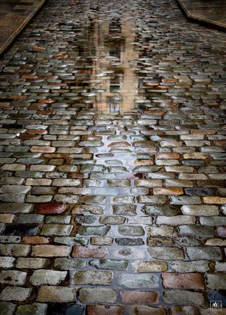 Color photo of a rain puddle in a colorful cobblestone street reflecting the facade of a yellow building.