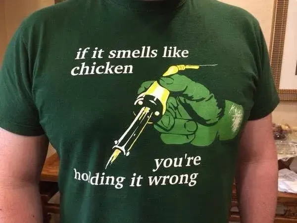 Photo of a green t-shirt with a hand holding a soldering iron and the text "if it smells like chicken you're holding it wrong"