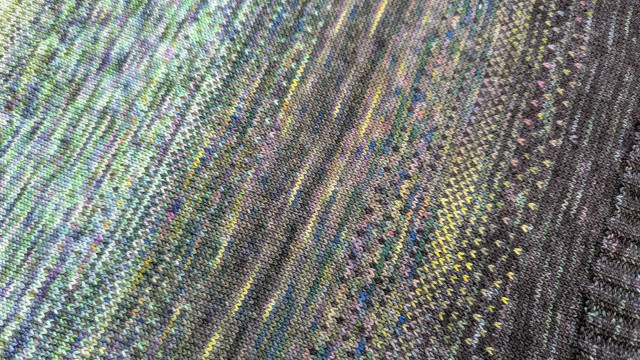 A closeup view of a piece of knitting. It has three shades of green yarn, lighter to darker, which fade into each other with some stranded colourwork that dithers the colours. Despite the sections where both yarns appear in the same rows, it still has sort of a striped appearance.