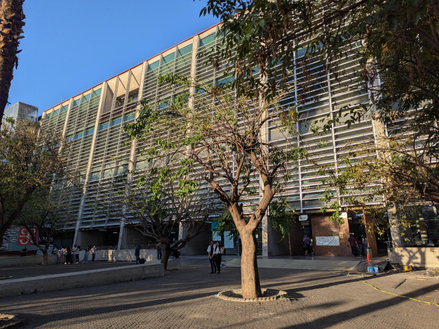 Outside of a wide 6 or 7 storey tall building from the outside. The facade has a lot of horizontal slats. Some small trees outside