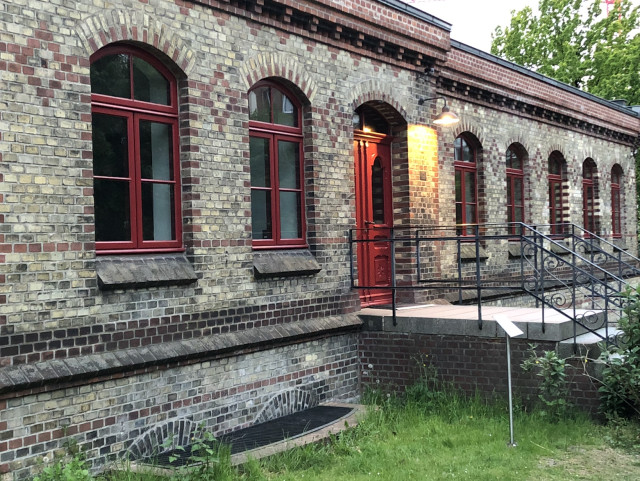 Langes altes Gebäude aus hellem Backstein mit gemauerten Mustern aus rotem Stein. Die Fensterreihe (nur eine Etage) hat passend dazu rote Rahmen. In der Mitte eine rote Tür die über kurzer Treppe zu erreichen ist. Neben der Treppe eine Infotafel.