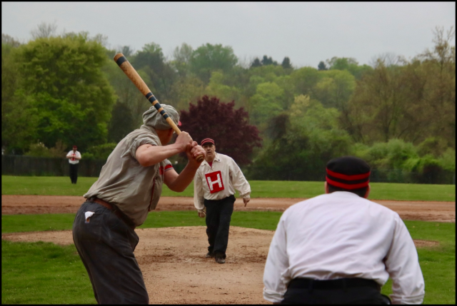 A pitcher, standing on a patch of dirt, pitches underhand to a batter near a catcher. In the background is a fielder standing on a grassy field, then many lush trees beyond. All the people are in old-timey clothes and hats; the pitcher's shirt has a big H on it. Photo from https://hobokennine.jimdofree.com/2026-photos-videos/