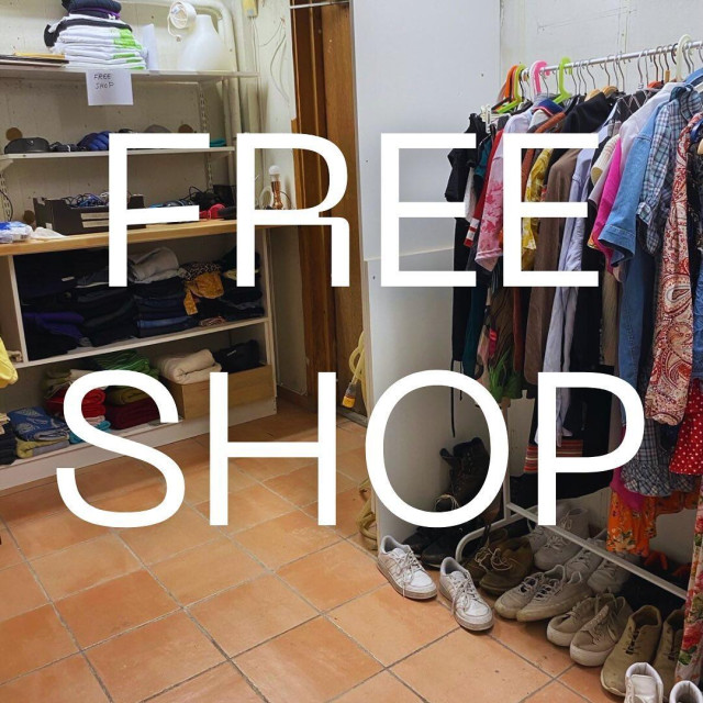 A small room with a tiled floor arranged as a "Free Shop," featuring shelves filled with folded clothes on the left, a rack of hanging clothing on the right, and rows of shoes lined up underneath. Large white text reading "FREE SHOP" is overlaid across the center.

