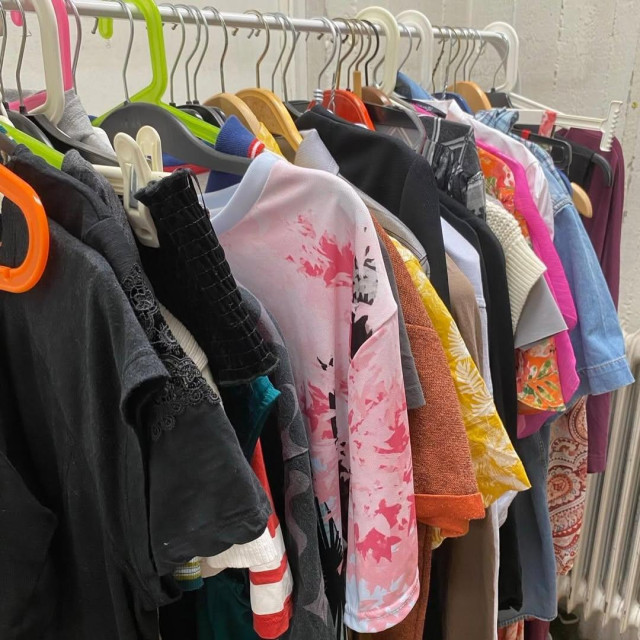A close-up view of a clothing rack tightly packed with a variety of colorful t-shirts, patterned shirts, and sweaters hanging on mismatched plastic and wooden hangers.

