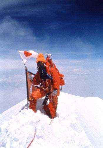 A climber in a thick orange suit standing on the snow-capped top of Mount Everest and holding onto a Japanese flag.