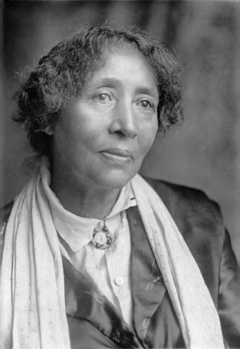 Black & white photo of a black woman, in maybe her fifties or early sixties?  Her face is unlined, but she *feels* old.  It is a formal portrait and she is looking 45 degrees off camera.  She is wearing a white or light-coloured blouse with a brooch at the neck, a darker blazer made of a shiny material, and a long, light-colour scarf.

Her mostly dark hair is short, and not crimped, exactly, but looks wiry & wavy.  Her expression seems a mixture of serious & maybe weary.  She does not look grim, but she certainly doesn't look happy either.