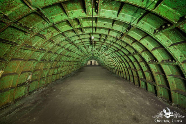 A long, arched green tunnel, constructed from ribbed metal panels, extends into the distance, revealing a Cold War-era atomic shelter. The floor is dark concrete.