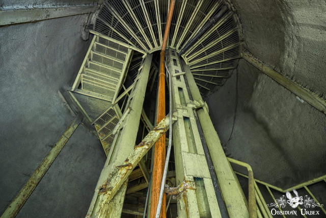 Looking up an abandoned spiral staircase inside the Rákosi Bunker. Rusting yellow-green metal stairs and orange pipes ascend towards a grated ceiling.