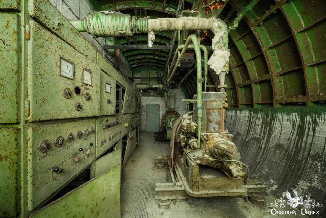 A decaying, green, subterranean bunker room with old machinery. Rusty control panels line the left, facing a large diesel generator on the right.