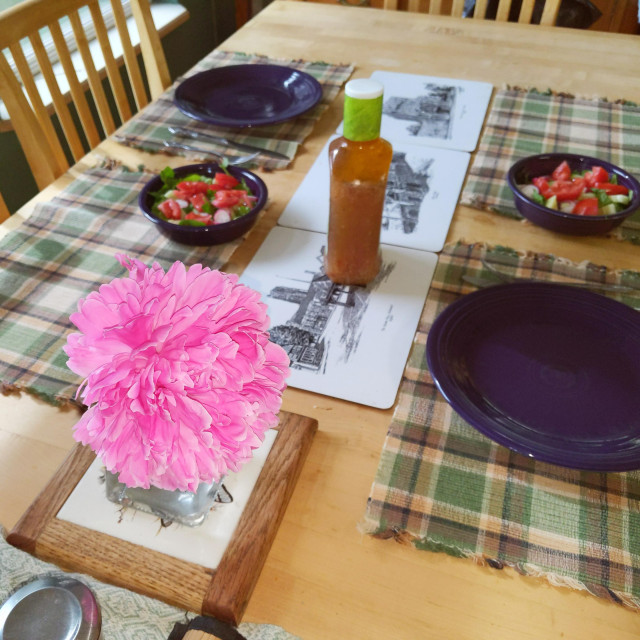 A very large pink #peonies #flower on a hard maple wood dining table. There are a couple of bowls of salad, empty plates, and the turned bottle of salad dressing as to not reveal the secret. 