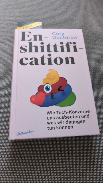 Das Buch Enshittification - Wie Tech-Konzerne uns ausbeuten und was wir dagegen tun können. Ein rosa Umschlag. Das Cover ziert ein buntes Scheißhaufen-Emoji das zwinkernd ein Herz küsst. 
Author: Cory Doctorow Verlag: Blümenbar