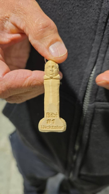 A hand holding a small 3D printed toy shaped like an erect penis with a man's head. It reads "Bibi #1 Dicktator" (referencing Israeli PM Benjamin "Bibi" Netanyahu).