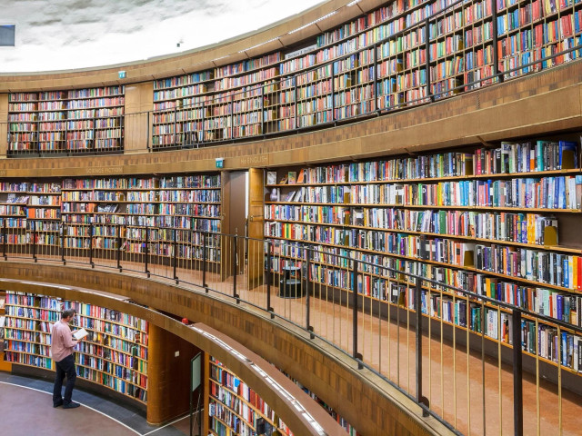 Photo of the interior of a 3-storey library, showing multiple rows of books. A man on the ground level is reading a book.