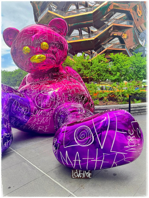 A color photo taken in front of the Hudson Yards building of a giant sized pink and purple metallic teddy bear. It is leaning back on its arms with its feet out so you can see the bottom of the foot. The bottom of the foot has the words "love matters" scratched into it in white. And the rest of the bear is covered with different words that have been scratched into it. I can see the words dream, desire and many others. The bear's eyes and nose are a bright yellow and behind it is a great deal of trees and shrubbery. Right above the trees can be seen a few floors of the Vessel, a sculpture that looks a bit like a beehive with different sets of stairs for people to walk up and see the view. It was a bright sunny day.