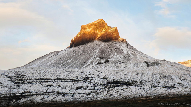 A colour photo of a mountain beneath a slightly cloudy sky. The flanks are snowy but ridges of dark rock have been scoured clear of ice by the winds. The shoulders of the mountain are at roughly 25 degrees to the horizontal, and the top is a volcanic plug with upright sides and a jagged top. Low sunlight from behind the viewpoint is being obscured by a range of mountains at the viewer's back, and only this craggy tip is sunlit.