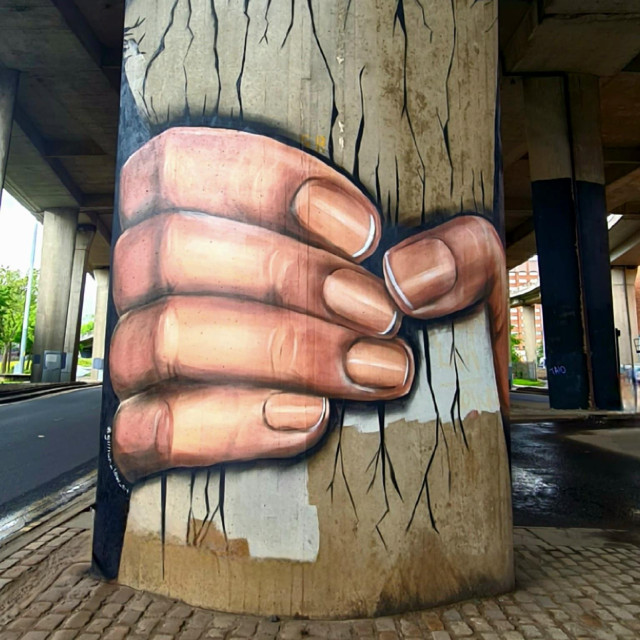A mural of a hand apparently crushing the pillar of a 1960s concrete motoway bridge.