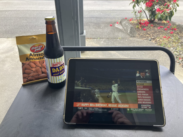 A small outdoor table sits beside a window or covered patio area on a rainy day. On the table are a bag of Dan-D Pak almonds, a glass bottle of Diet Dad’s Old Fashioned Root Beer, and a tablet in a protective case. The tablet is playing a sports broadcast showing old footage of Reggie Jackson in a New York Yankees uniform, with a banner reading “Happy 80th Birthday, Reggie Jackson!” and sports headlines running down the side of the screen.

The pavement outside looks wet from recent rain. In the background, a flowering shrub with bright pink blossoms sits beside the sidewalk, with scattered petals on the ground.