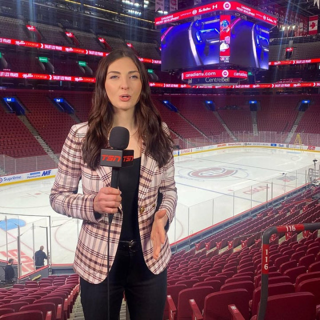 A sports reporter stands in the seating area of an empty hockey arena holding a TSN microphone, speaking toward the camera. She wears a light plaid blazer over a black top and black pants. Behind her is the ice surface at the Bell Centre, home of the Montreal Canadiens, with rows of red seats, rink boards, and a large overhead scoreboard displaying Canadiens branding and sponsor graphics. Bright arena lighting illuminates the scene.