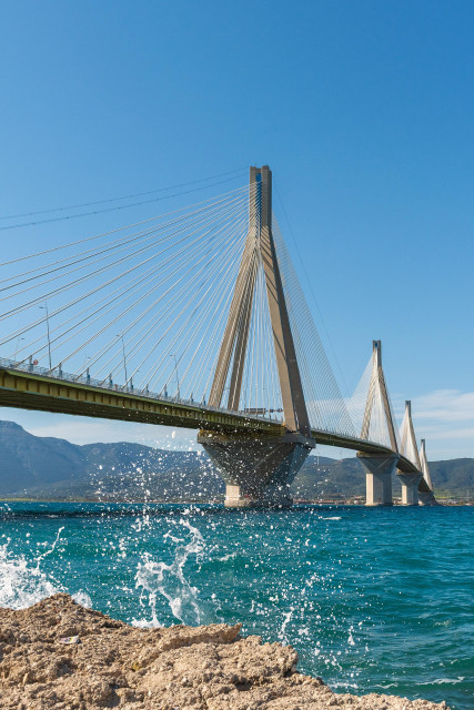 Das Bild zeigt eine beeindruckende Schrägseilbrücke, die sich majestätisch über das türkisfarbene Meer erstreckt, während Gischt aufspritzt.

Das Bild zeigt eine beeindruckende Schrägseilbrücke an einem sonnigen Tag, deren schlanke Pylone und zahlreichen Stahlseile sich gegen einen leuchtend blauen Himmel abzeichnen. Die Brücke überspannt ein tiefblaues Meer, auf dem sich weiße Gischtfontänen erheben, die durch Wellen oder den Wind ausgelöst wurden und wie kleine Diamanten funkeln. Im Vordergrund sind felsige Uferbereiche zu sehen, die mit braunen und sandfarbenen Steinen bedeckt sind. Im Hintergrund erheben sich sanfte, grüne Berge, die von dem klaren, blauen Himmel eingerahmt werden.