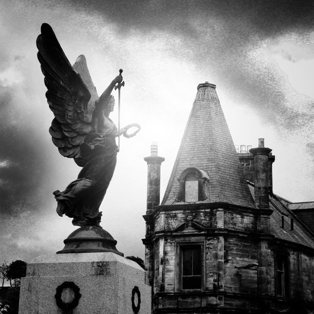 Atmospheric black and white photo of Lockerbie War Memorial, a statue of winged victory holding a sword and wreath, on a square plinth. Behind the memorial are the late 19th century Tower Buildings, with dark clouds above.
