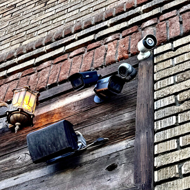 Several security cameras mounted to an exterior wall pointing in various directions. The wall itself is made of a variety of textures such as weathered wood and at least three different types of brick.