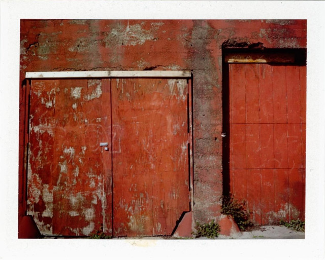 A red-brown concrete wall set in with wooden doors in various states of decay. The overall feel is of an abstract expressionist painting. 