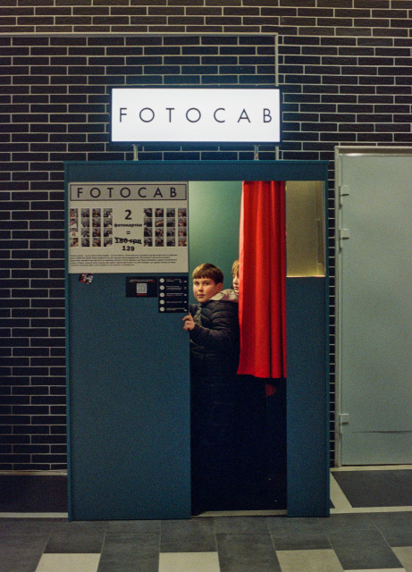 Two children peeking out from behind a red curtain inside a photo booth called "FOTOCAB", set against a dark brick wall. The booth is teal-colored with a illuminated sign above and a price list panel on the front