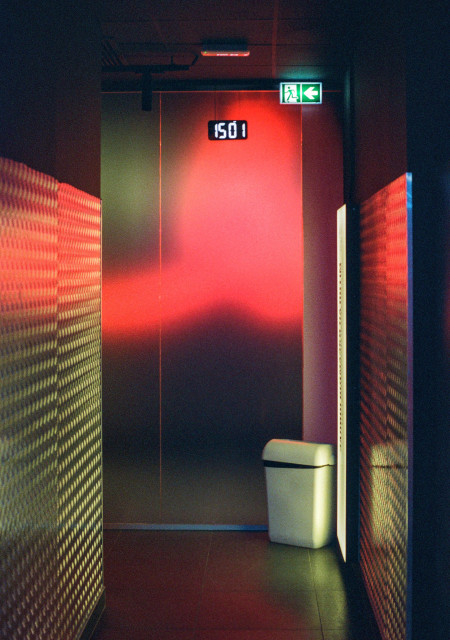 A dim, empty interior space with a metallic wall at the far end, washed in intense red light. Above it, a digital clock reads 15:01, while a green emergency exit sign glows nearby. Textured illuminated panels on both sides create a strange, almost futuristic corridor effect. A small pale trash bin stands near a bright doorway, adding an ordinary detail to the otherwise cinematic, uneasy atmosphere.