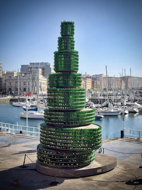 Scultura di bottiglie verdi impilate a forma di albero, vicino a un porto con barche e edifici sullo sfondo.
