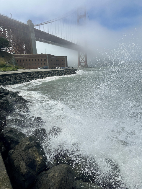 The Golden Gate Bridge and Fort Point National Historic Site. Fog obscures the bridge in the background, but the brick fort stands clearly on the road by the coastline. A large wave of sea water splashes the rocky coast (and the photographer.)